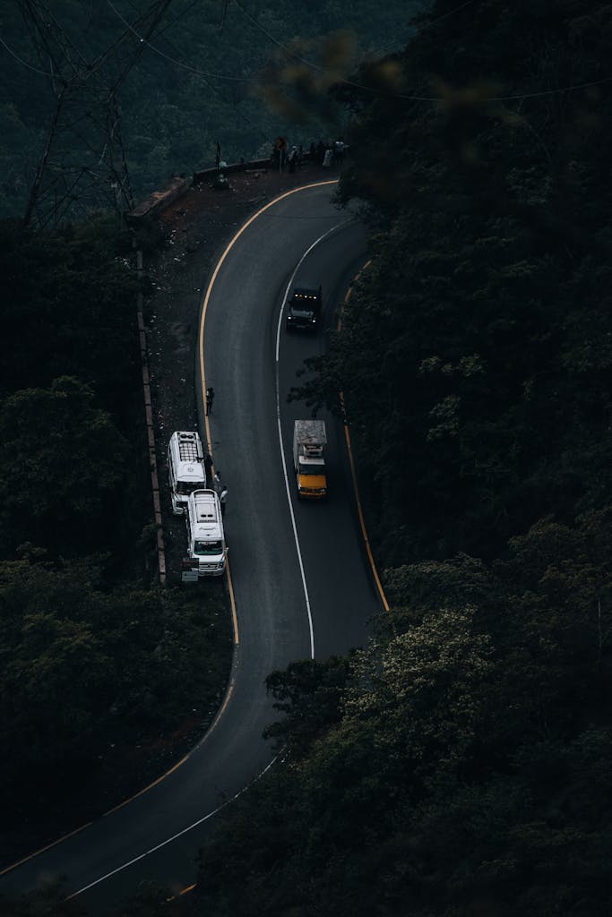 An aerial shot of a curved mountain road with trucks navigating through dense forest on a misty day.